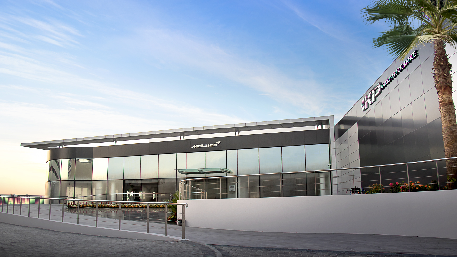 A photograph of a modern McLaren office. The two-story structure features a large, glass facade on the ground floor and a black panel with the white McLaren logo on the first.