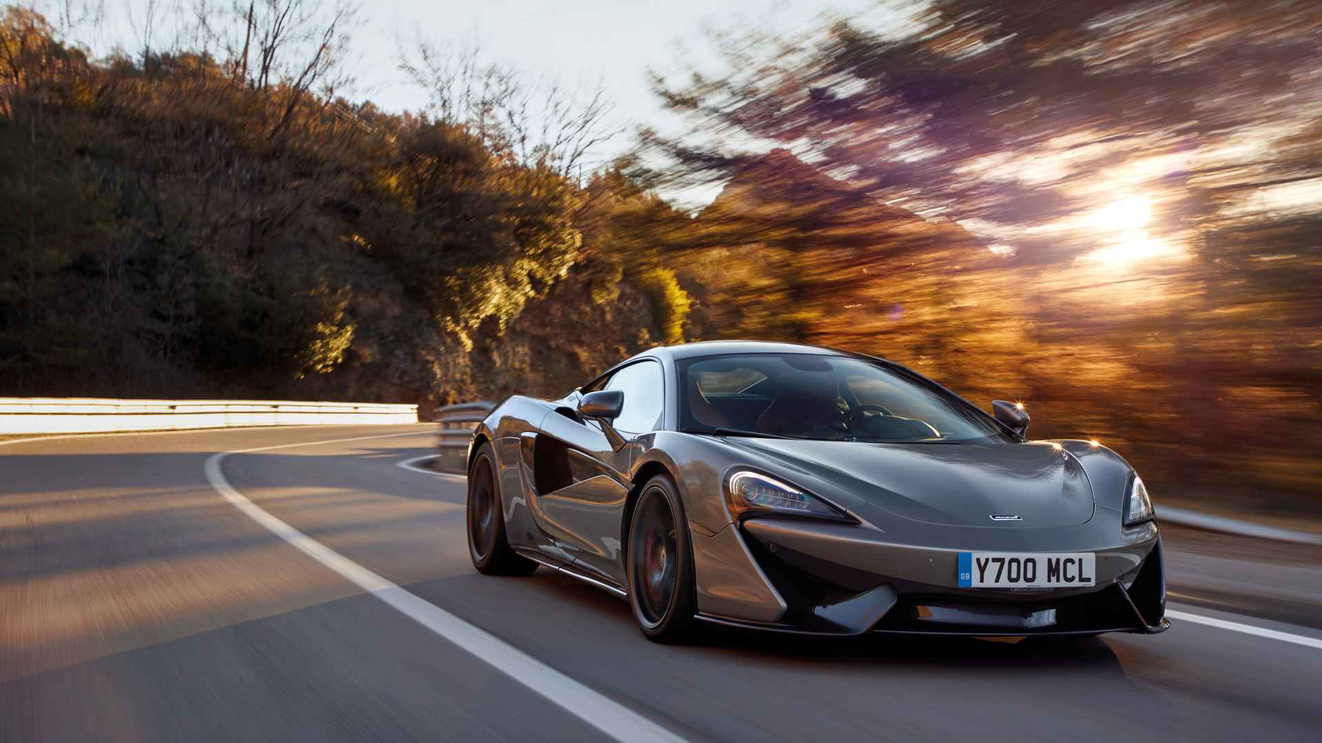 A grey McLaren 570S sports car is shown from a dynamic front-three-quarter view, driving on a winding road with sunlight filtering through the trees, creating a sense of speed and motion.