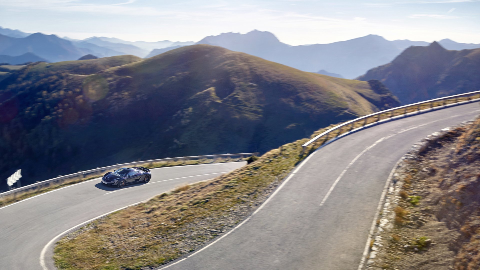 A panoramic shot of a grey McLaren sports car driving on a winding road with sharp turns through a majestic mountain landscape at sunset.
