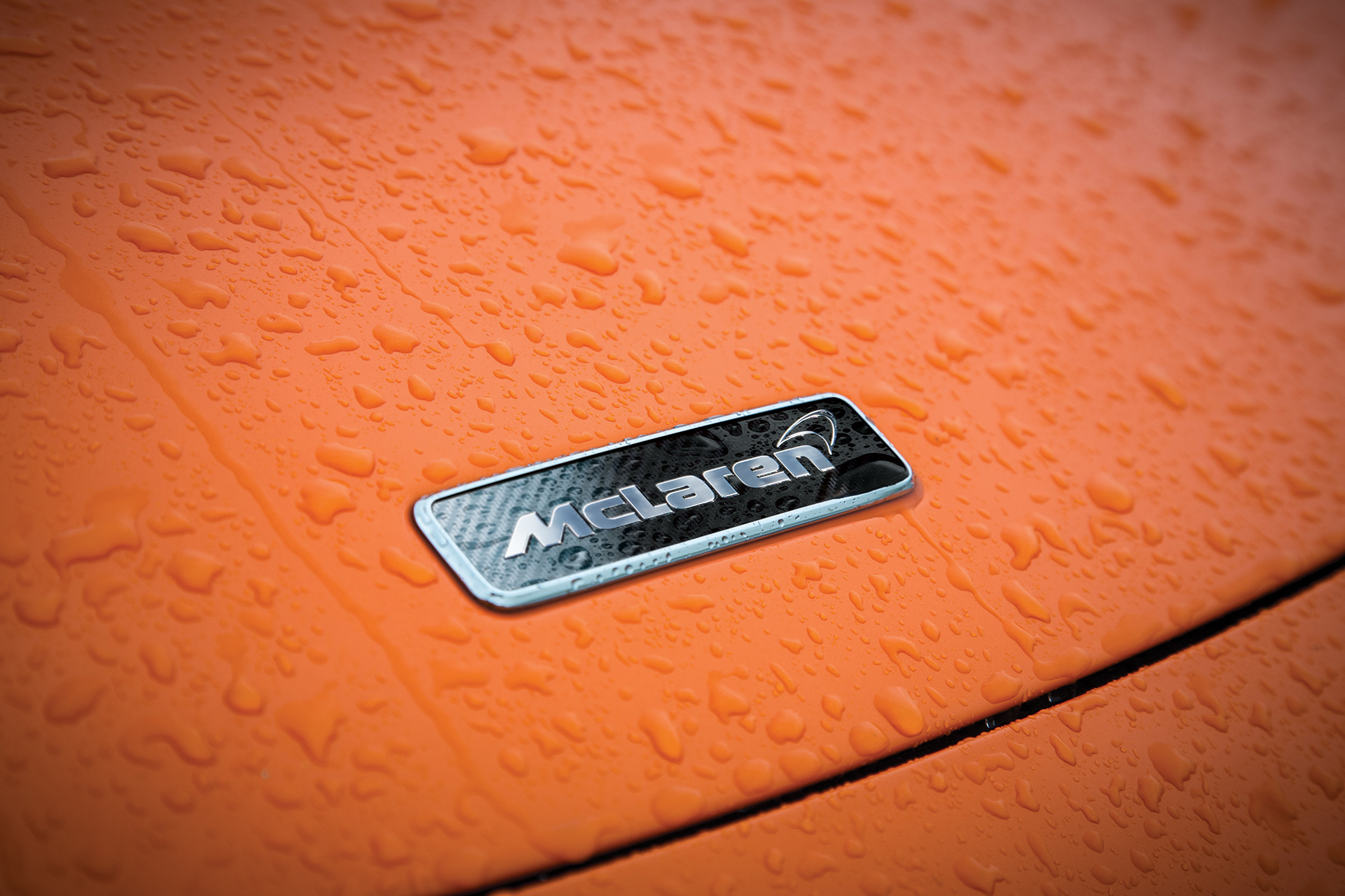 A close-up of the McLaren badge on the hood of an orange car, with water droplets on the surface, creating a wet and textured look.