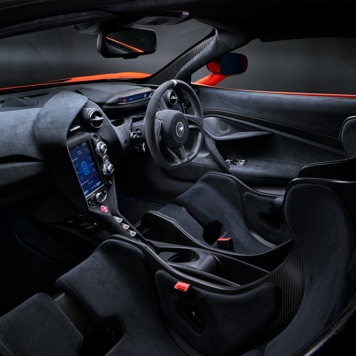 A detailed view of the interior of a McLaren 765LT, featuring a black Alcantara dashboard and racing seats, a digital infotainment screen, and carbon fiber trim.