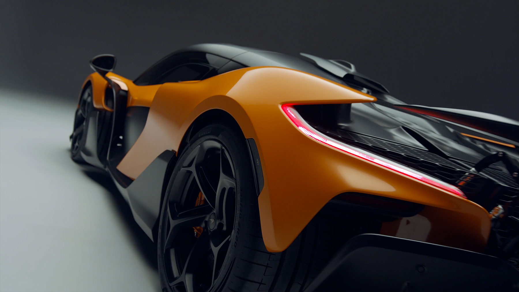  A dynamic low-angle, rear three-quarter shot of an orange and black McLaren supercar, highlighting its sleek body lines, intricate taillight design, and rear wheel.