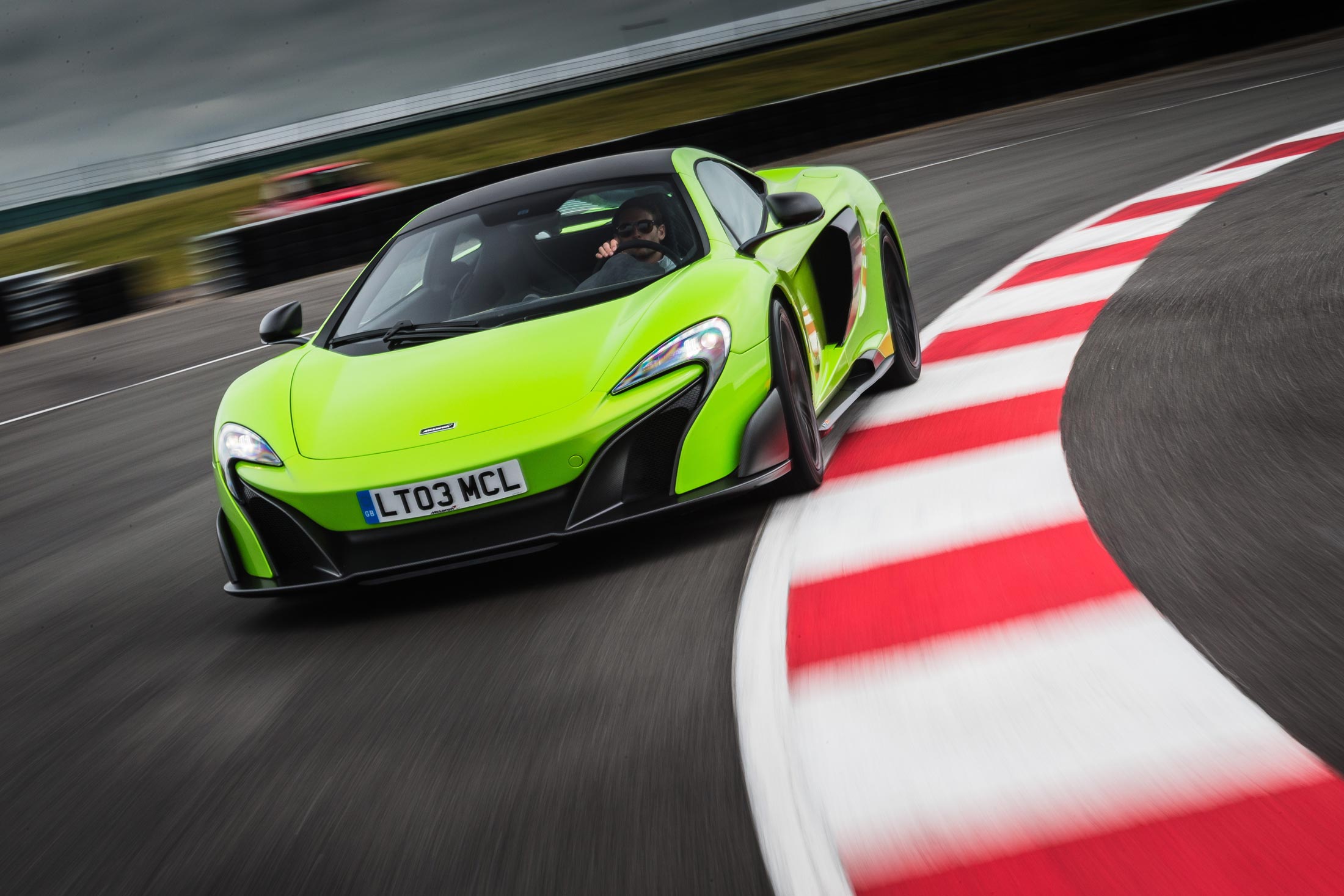 A dynamic front-three-quarter view of a lime green McLaren 675LT as it takes a sharp turn on a racetrack, with a red and white curb in the foreground.