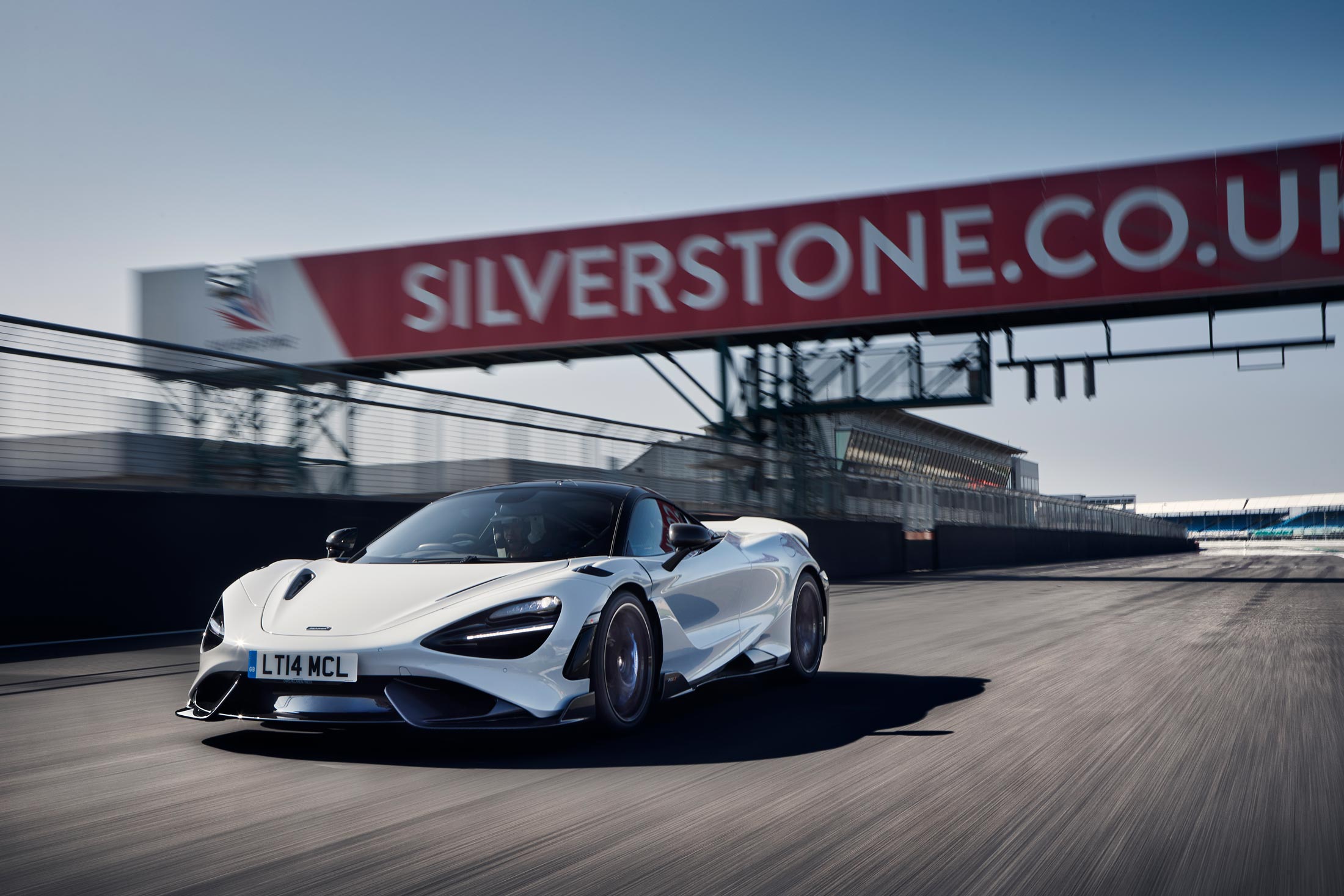 A dynamic front-three-quarter view of a white McLaren 765LT speeding down the main straight at the Silverstone racetrack, with the famous grandstands and bridge in the background.