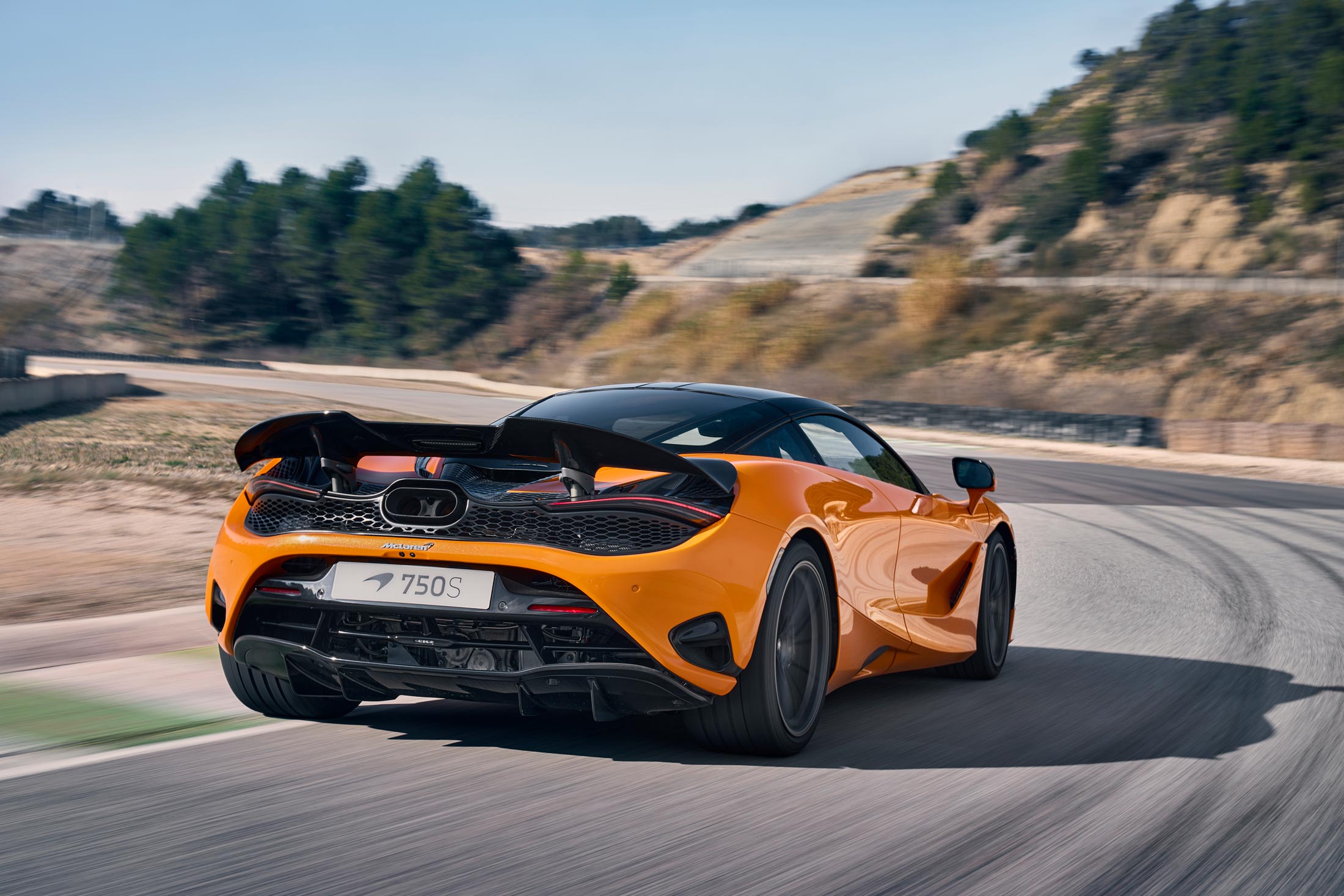 A rear-three-quarter view of a bright orange McLaren 750S on a winding racetrack, showcasing its aggressive rear spoiler, diffuser, and taillights.