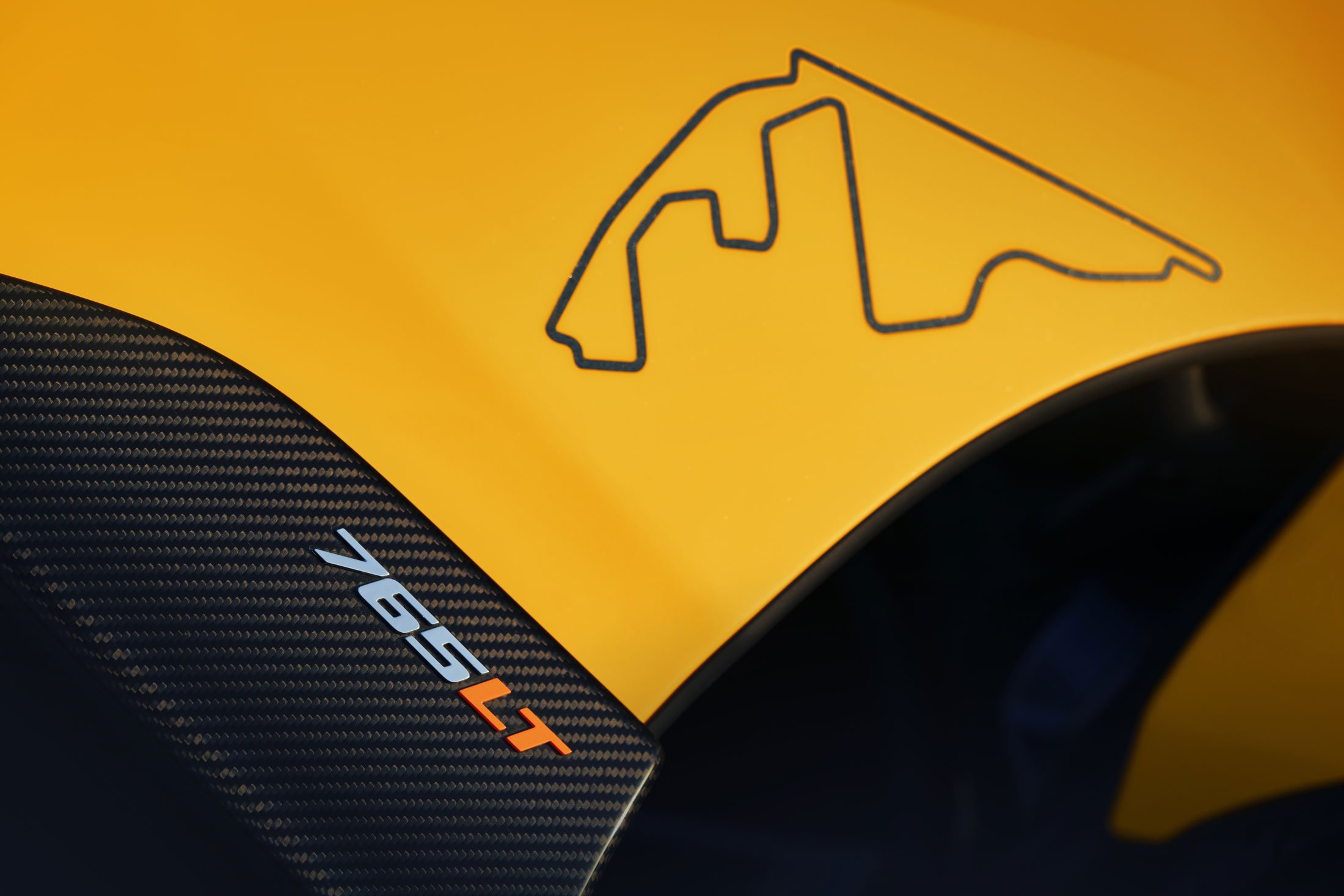A detail shot of a yellow McLaren 765LT's carbon fiber bodywork, with the '765LT' logo and a hand-drawn racetrack map on the hood.