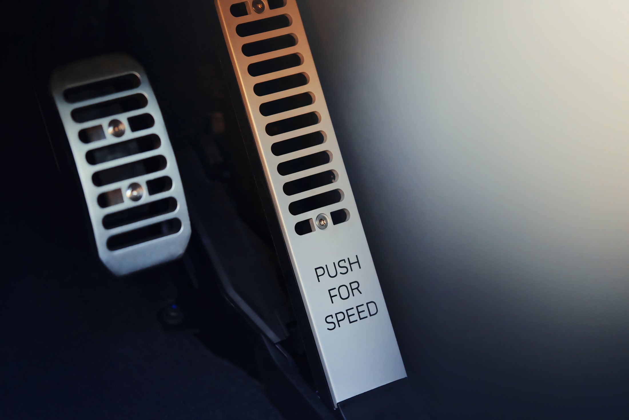 A close-up of a McLaren's metallic accelerator pedal, with the text 'PUSH FOR SPEED' engraved on it, alongside the brake pedal.
