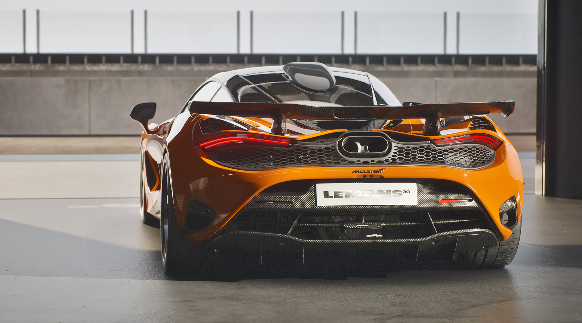 A rear-three-quarter view of an orange McLaren 750S Le Mans edition, highlighting its large rear spoiler, central exhaust, and the 'Le Mans 30' license plate.