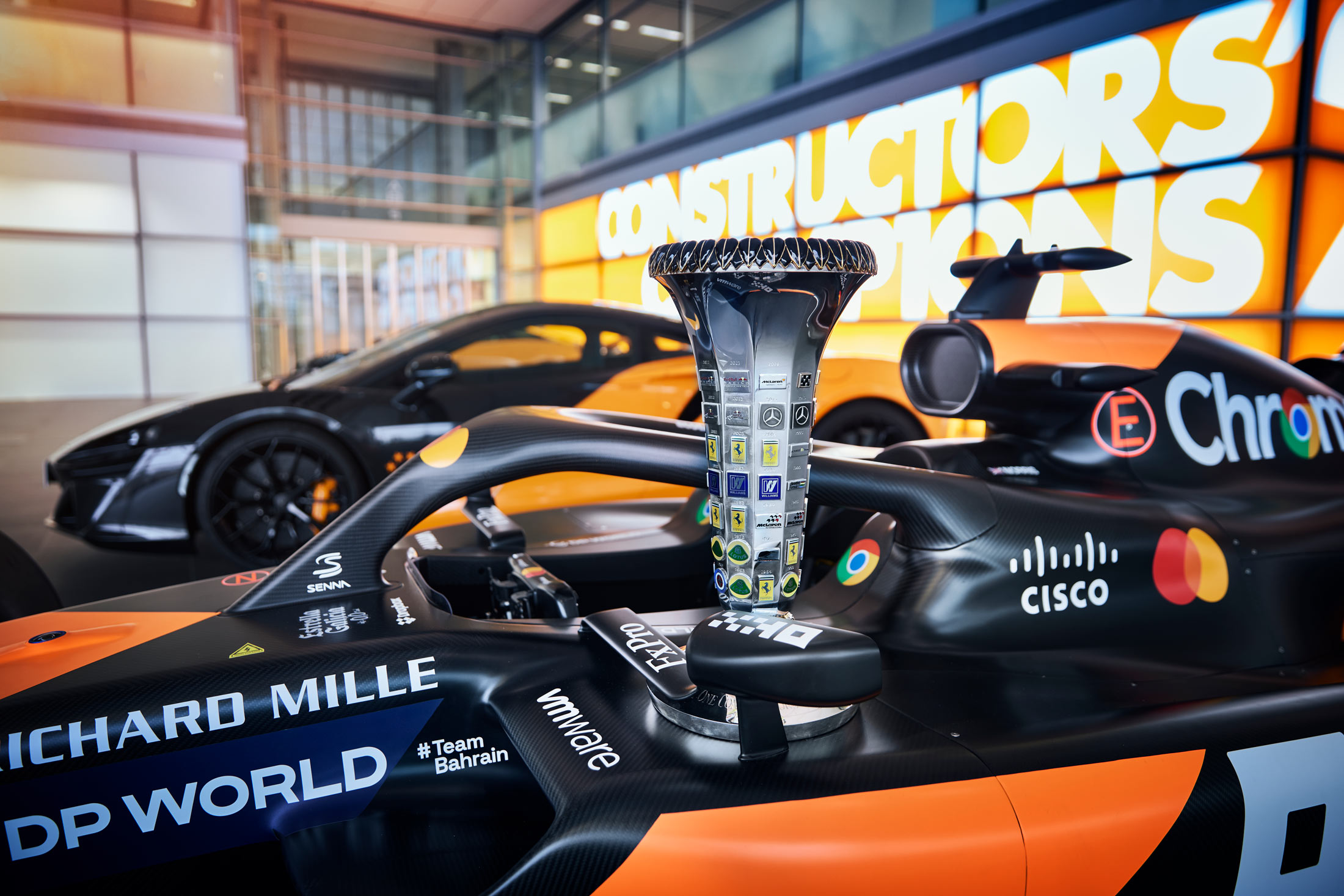 A close-up of a McLaren Formula 1 car's cockpit, with a large trophy placed on top of its chassis, and two road cars visible in the background.