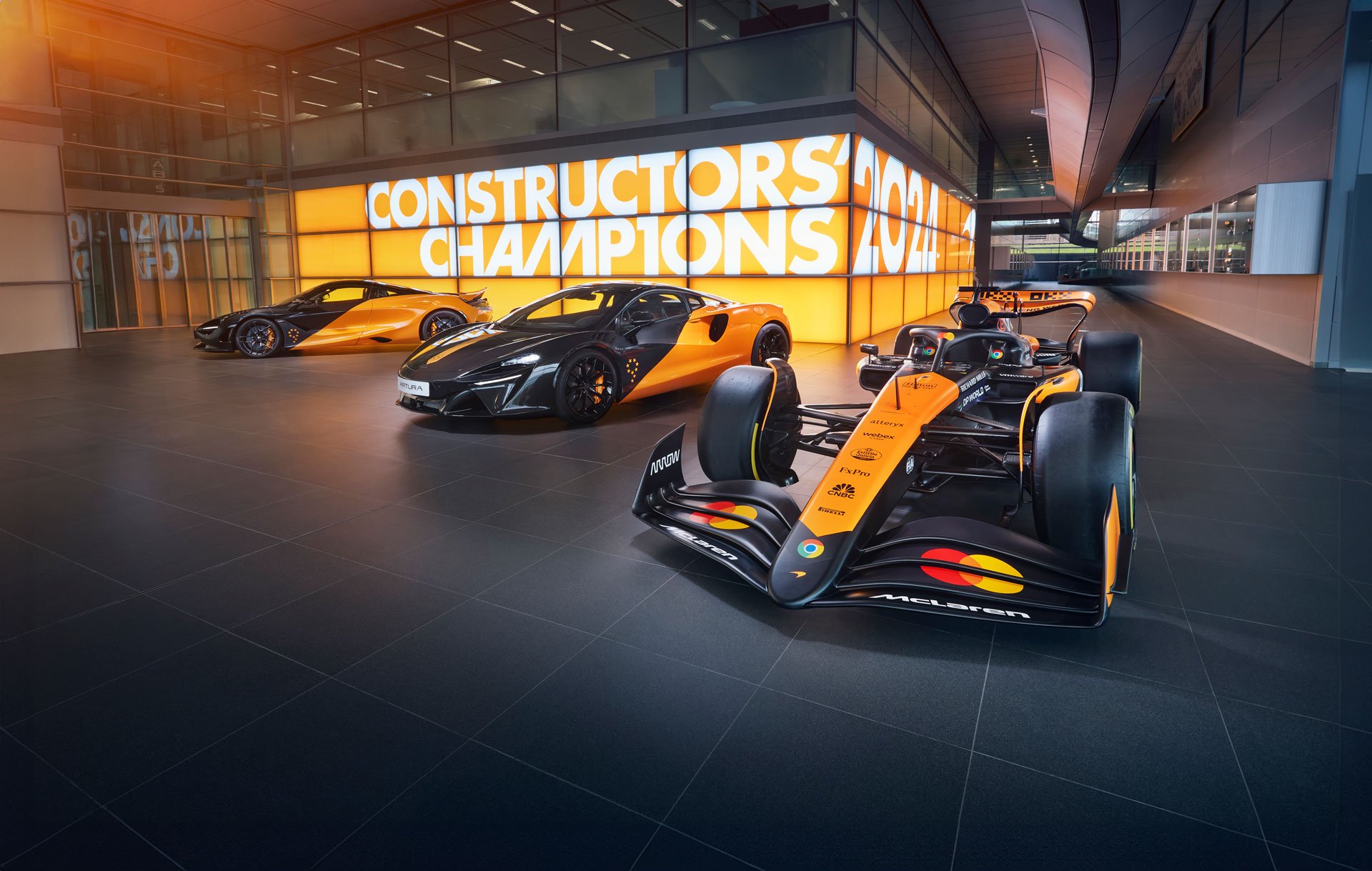 A hero shot of a McLaren Formula 1 car, a McLaren Artura, and another McLaren supercar, all in the iconic orange and black livery, displayed in a showroom.
