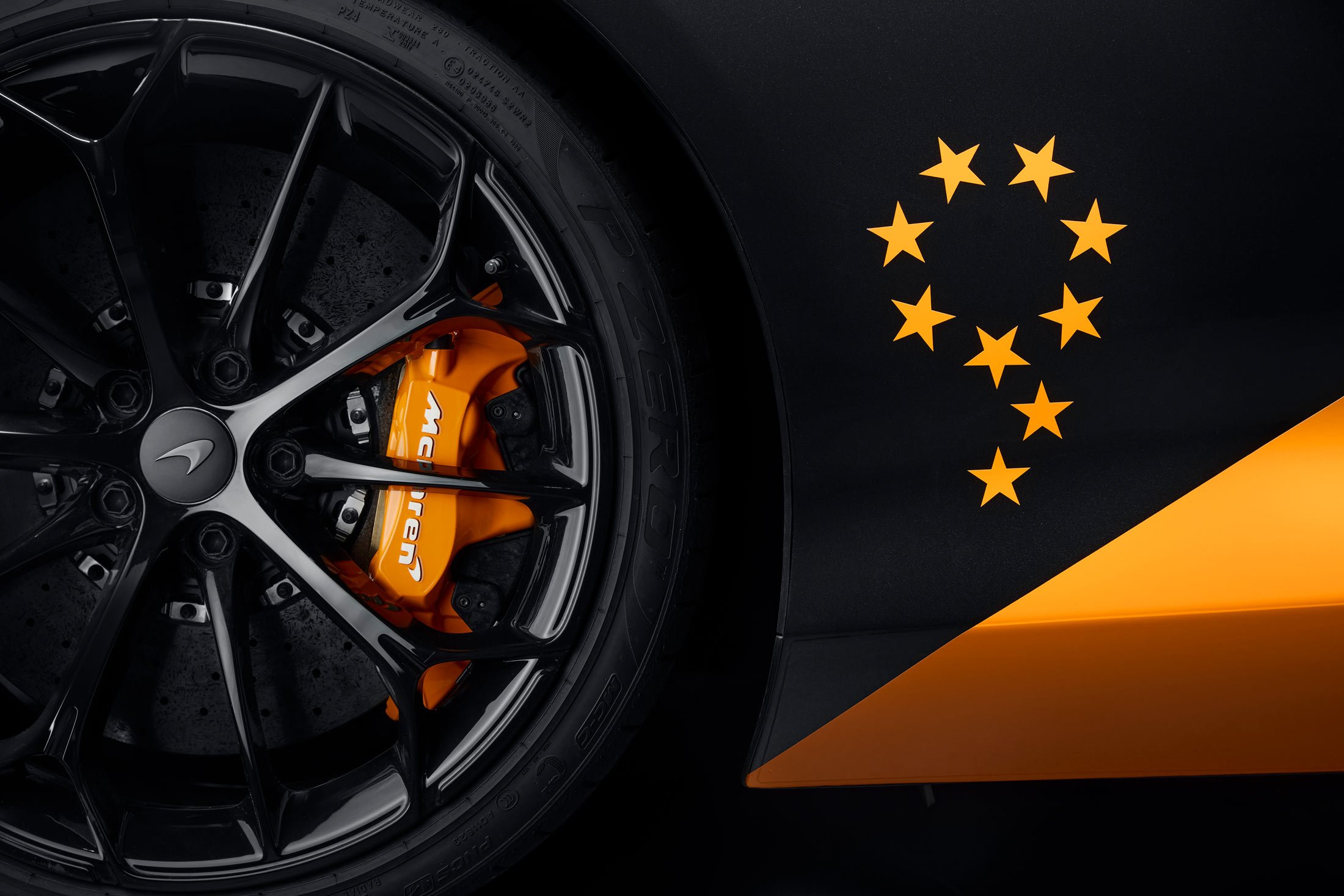 A close-up of a McLaren's front wheel, highlighting the orange brake caliper and a unique graphic of nine stars on the car's black bodywork.