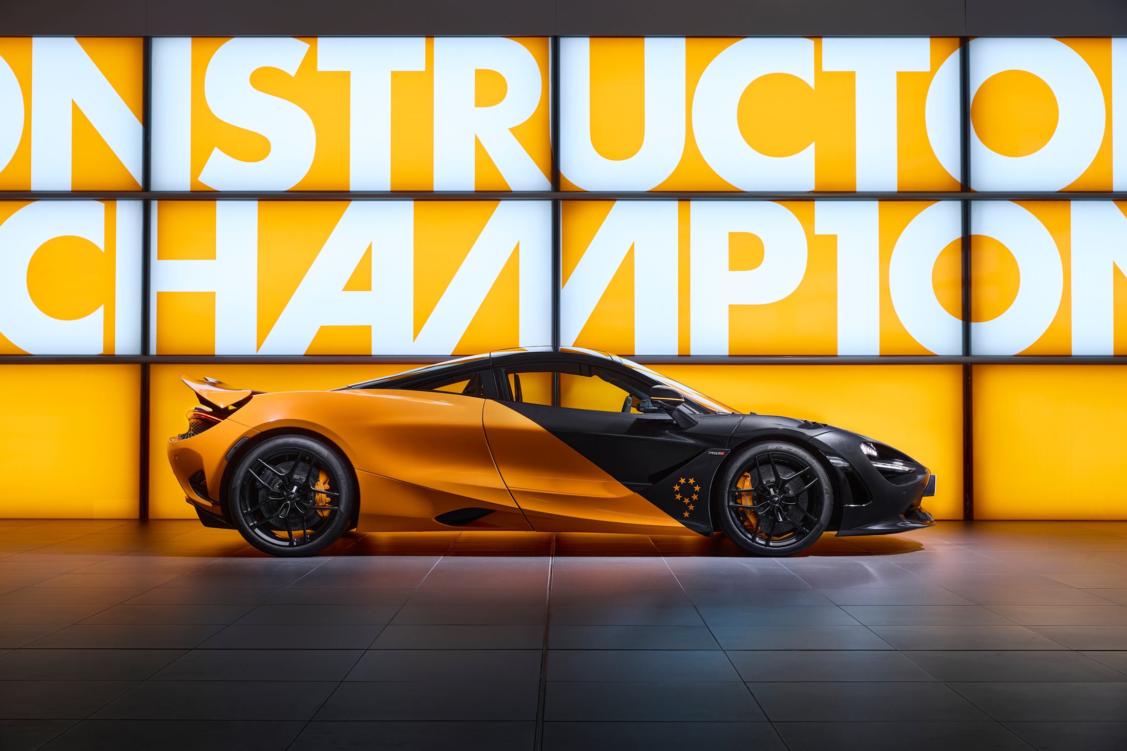 A side profile shot of a two-tone black and orange McLaren 720S supercar, with its livery celebrating McLaren's racing heritage, parked in front of a 'CONSTRUCTORS' CHAMPIONS' wall.