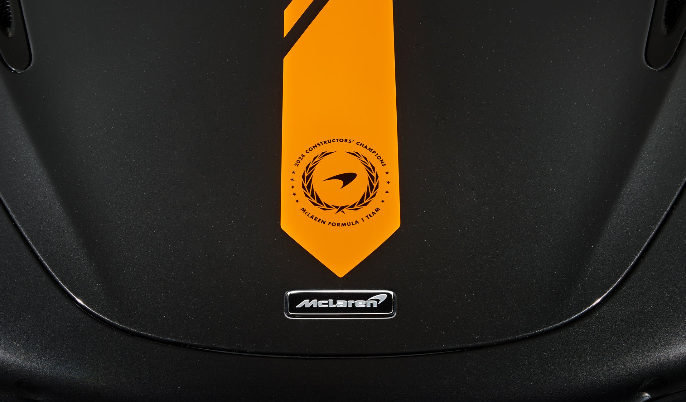 A close-up of a McLaren's hood, showcasing a commemorative badge celebrating the 1998 Formula 1 Constructors' Championship on a black and orange livery.