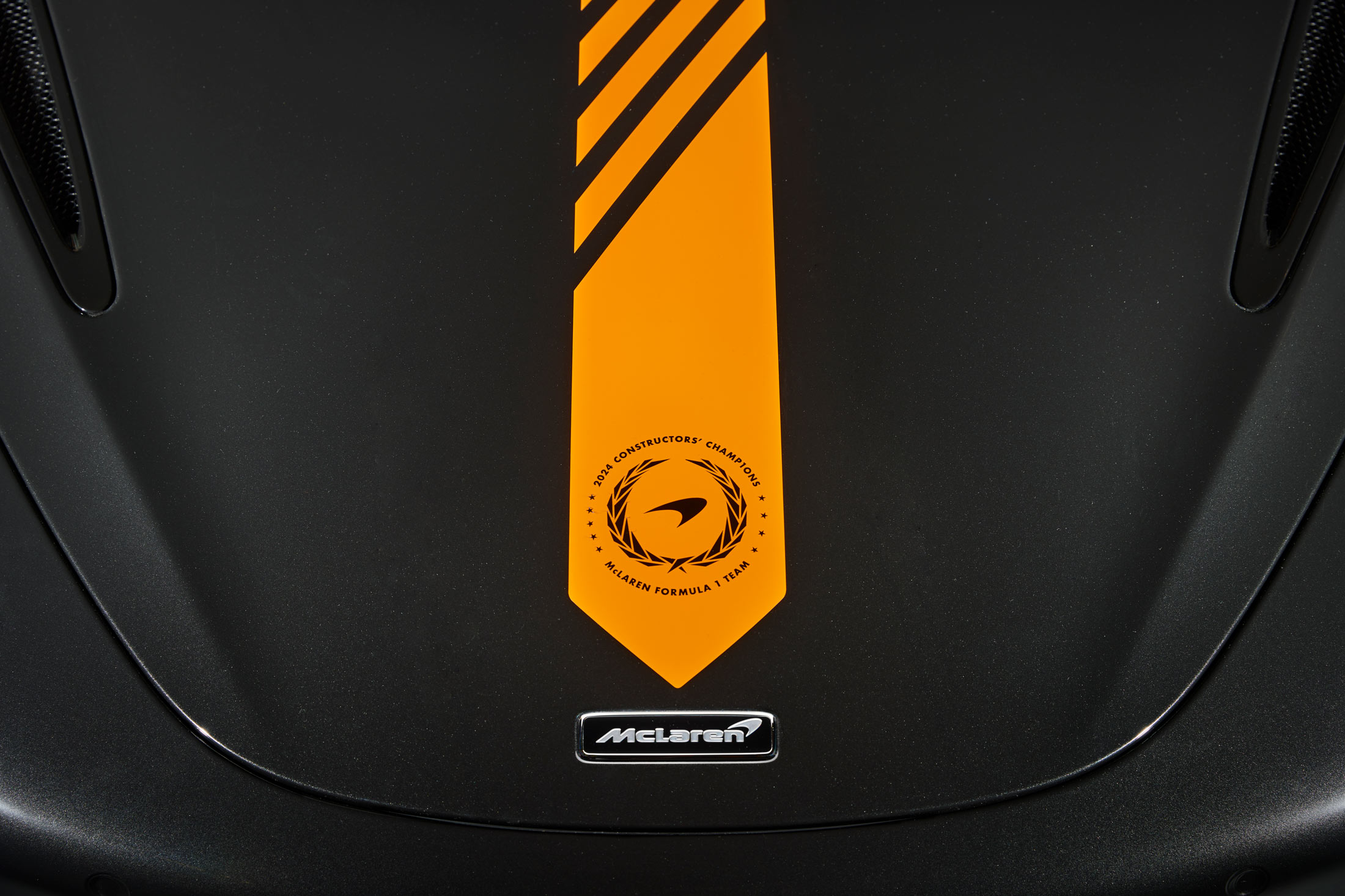 A close-up of a McLaren's matte black hood, showcasing an orange racing stripe with a badge that commemorates the 1998 Formula 1 Constructors' Championship.