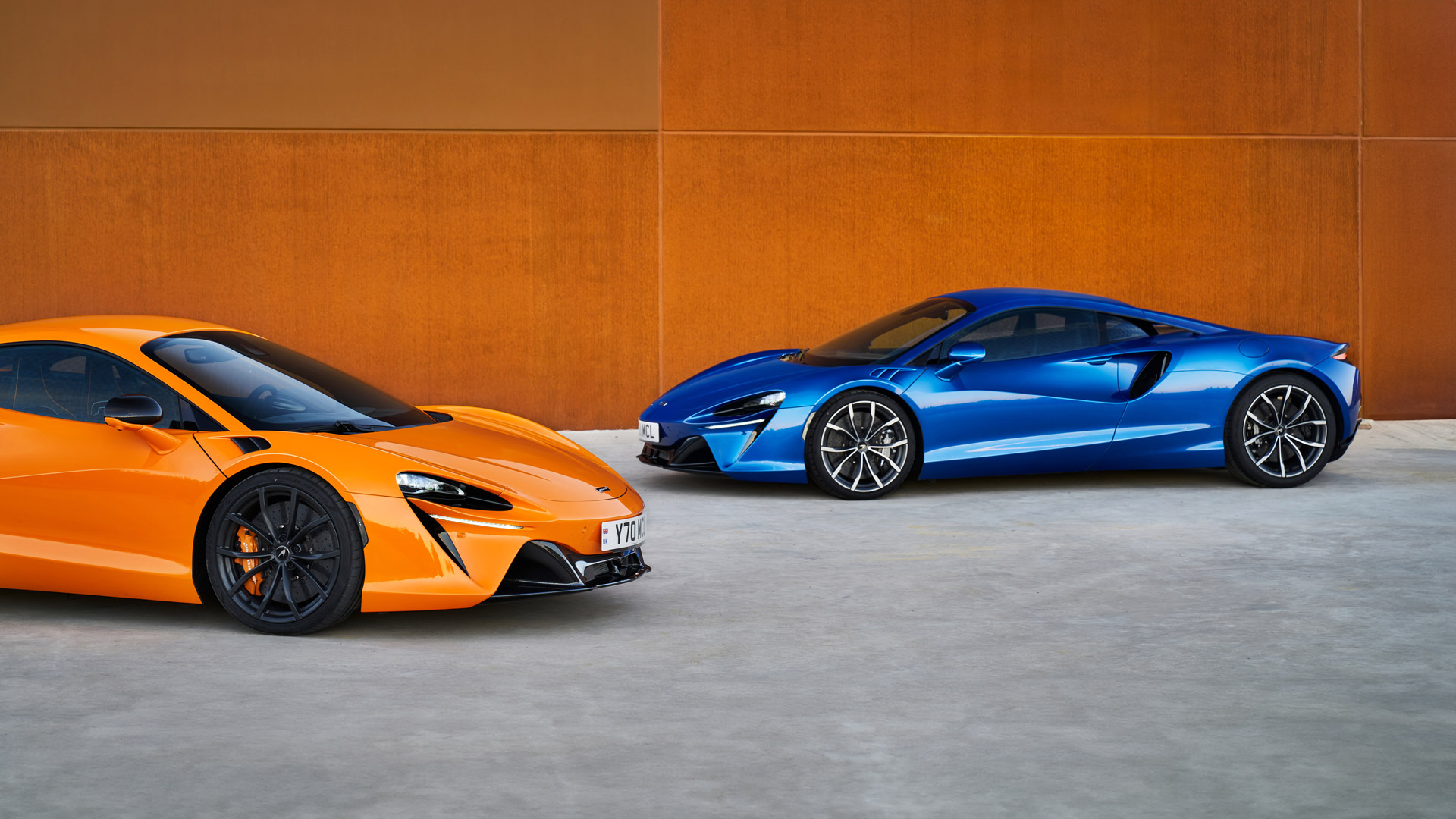 A side-by-side view of two McLaren Artura hybrid supercars, one in signature McLaren Orange and the other in vibrant blue, parked together.
