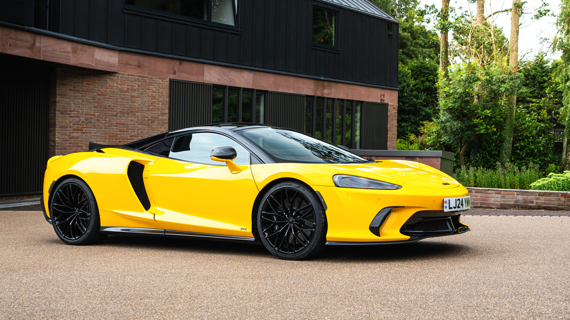 The bright yellow McLaren GTS grand tourer, with a black roof and wheels, parked in front of a modern brick and timber house.
