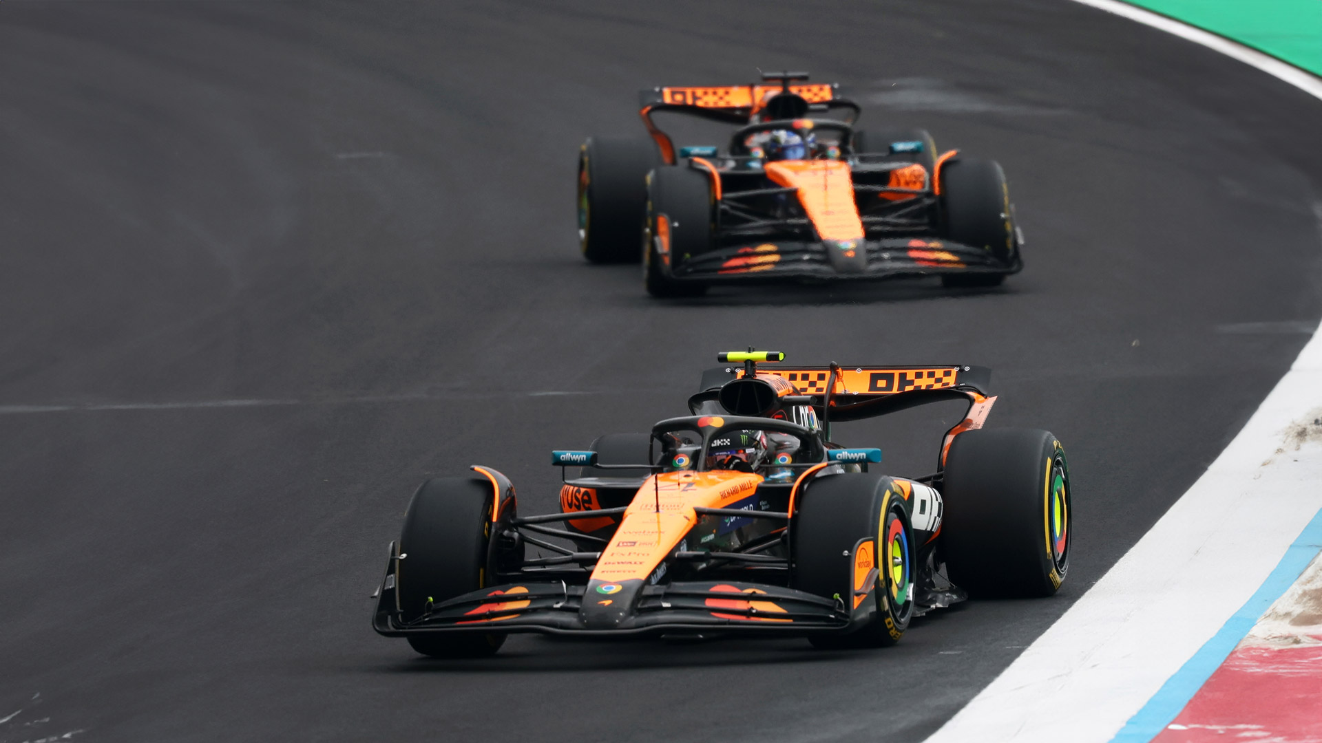 Two McLaren F1 cars, the MCL39, racing in formation on the circuit, highlighting the team's iconic papaya and black livery.