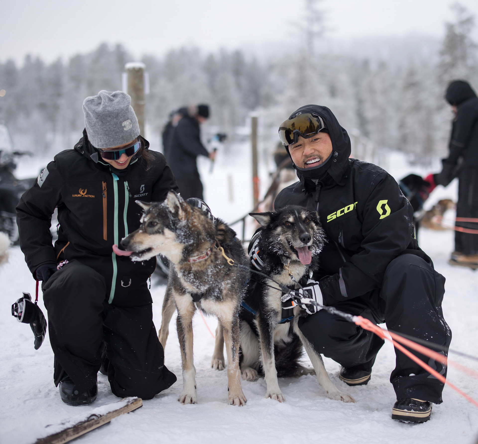 Two people kneeling in the snow with husky dogs, representing the non-driving activities available during a McLaren Arctic experience.