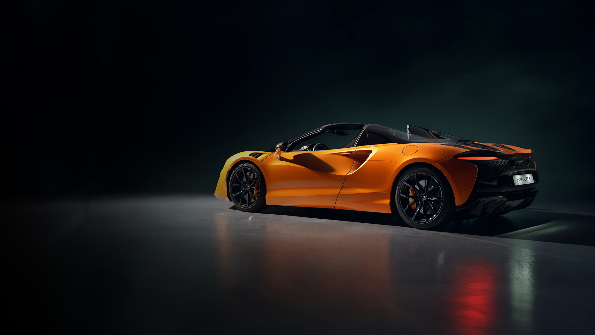 The breathtaking McLaren Artura Spider hybrid supercar in vibrant Volcanic Papaya Orange. A rear-quarter view highlights the convertible's aerodynamic profile and dramatic lighting in a studio, showcasing pure, electrified high-performance design.