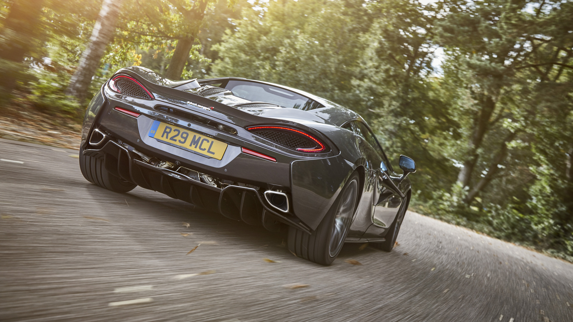 A rear low-angle shot of a dark grey McLaren supercar accelerating on a winding road, showcasing its distinctive taillights, exhaust pipes, and license plate, with sun-dappled trees in the background.