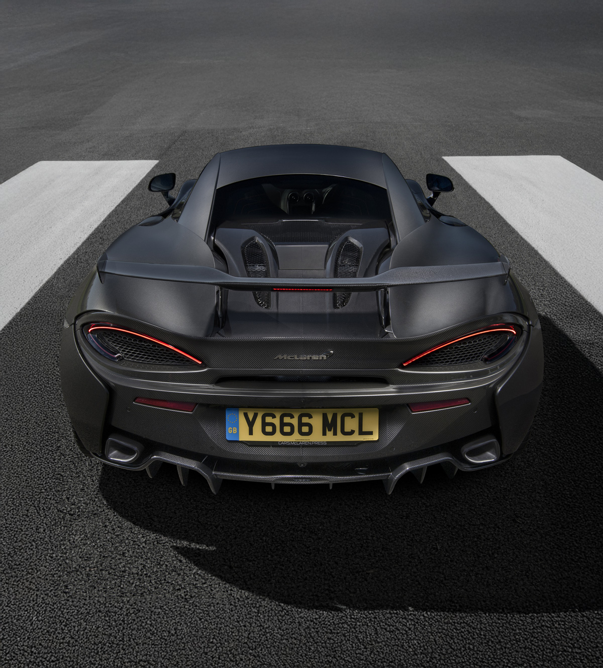 A high-angle, direct rear view of a dark grey McLaren 600LT, showcasing its distinctive exhaust pipes exiting through the top of the rear deck, aggressive diffuser, and prominent taillights on a racetrack.