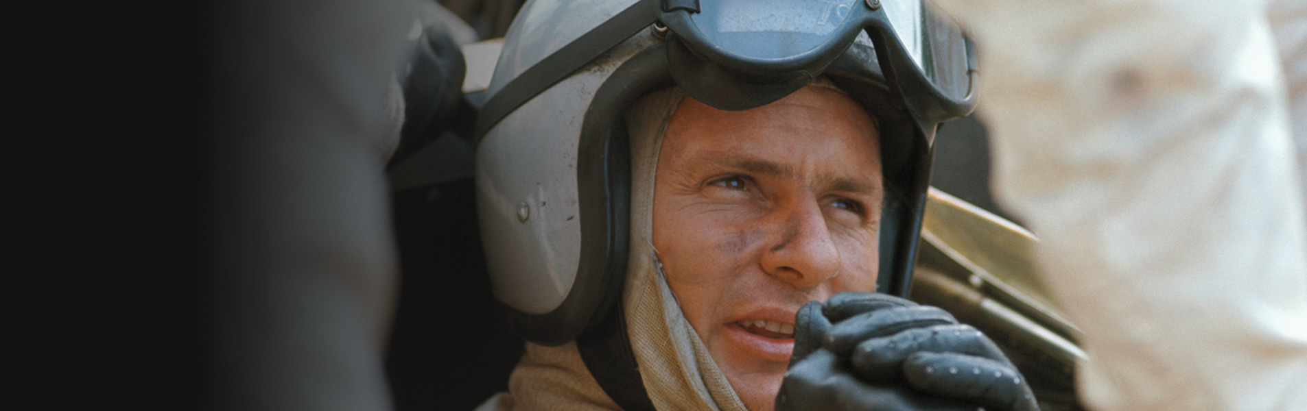 Close up of Bruce McLaren sat in a race car, wearing a helmet and driving gloves.
