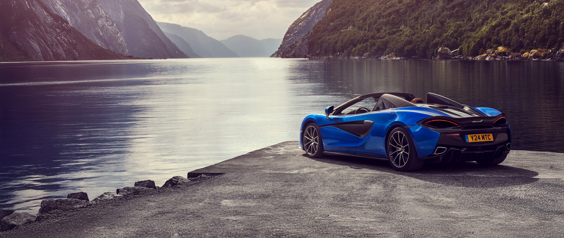 A blue McLaren 570S Spider convertible is parked by a scenic lake with a large mountain or cliffside in the background, showcasing its side profile.