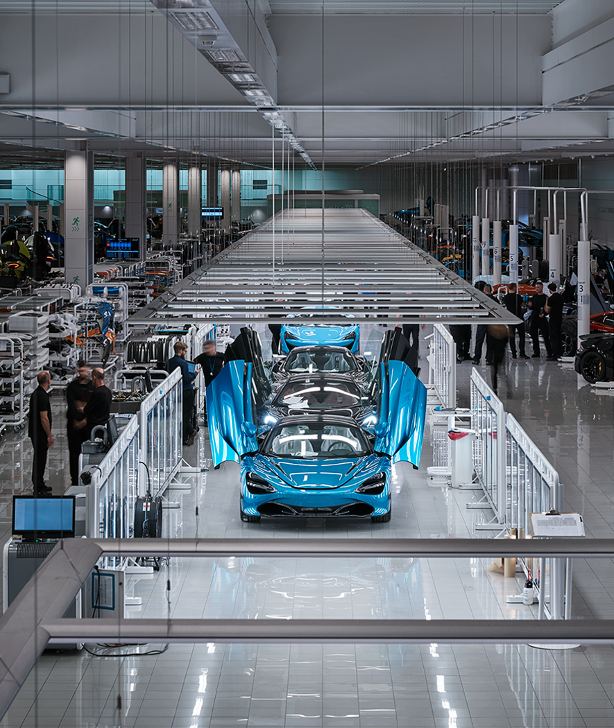 A high-angle view of the assembly line at the McLaren Production Centre. Several bright blue McLaren sports cars are lined up, with the closest one having its butterfly doors open.