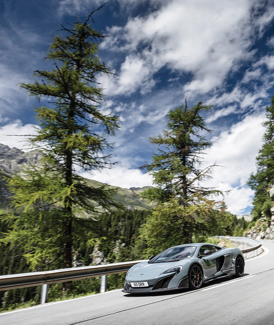 Grey McLaren 675LT driving on a mountainside road surrounded by trees.