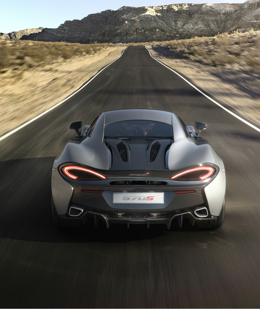 The rear of a grey McLaren 570S supercar with its taillights on, driving at high speed on a road in a desert landscape.