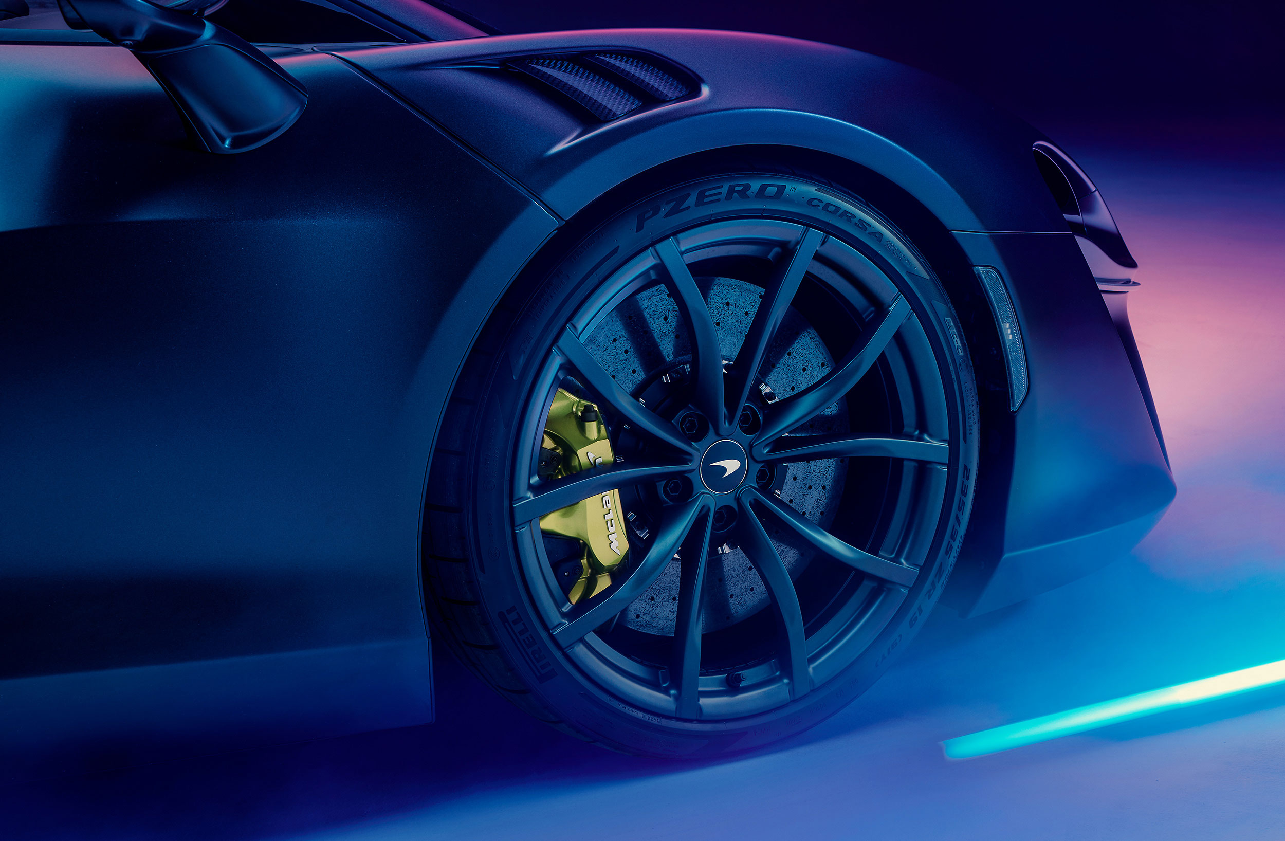 Close up of a Pirelli tyre on a McLaren wheel with a lime green caliper. The car is lit up by blue and pink neon lights.