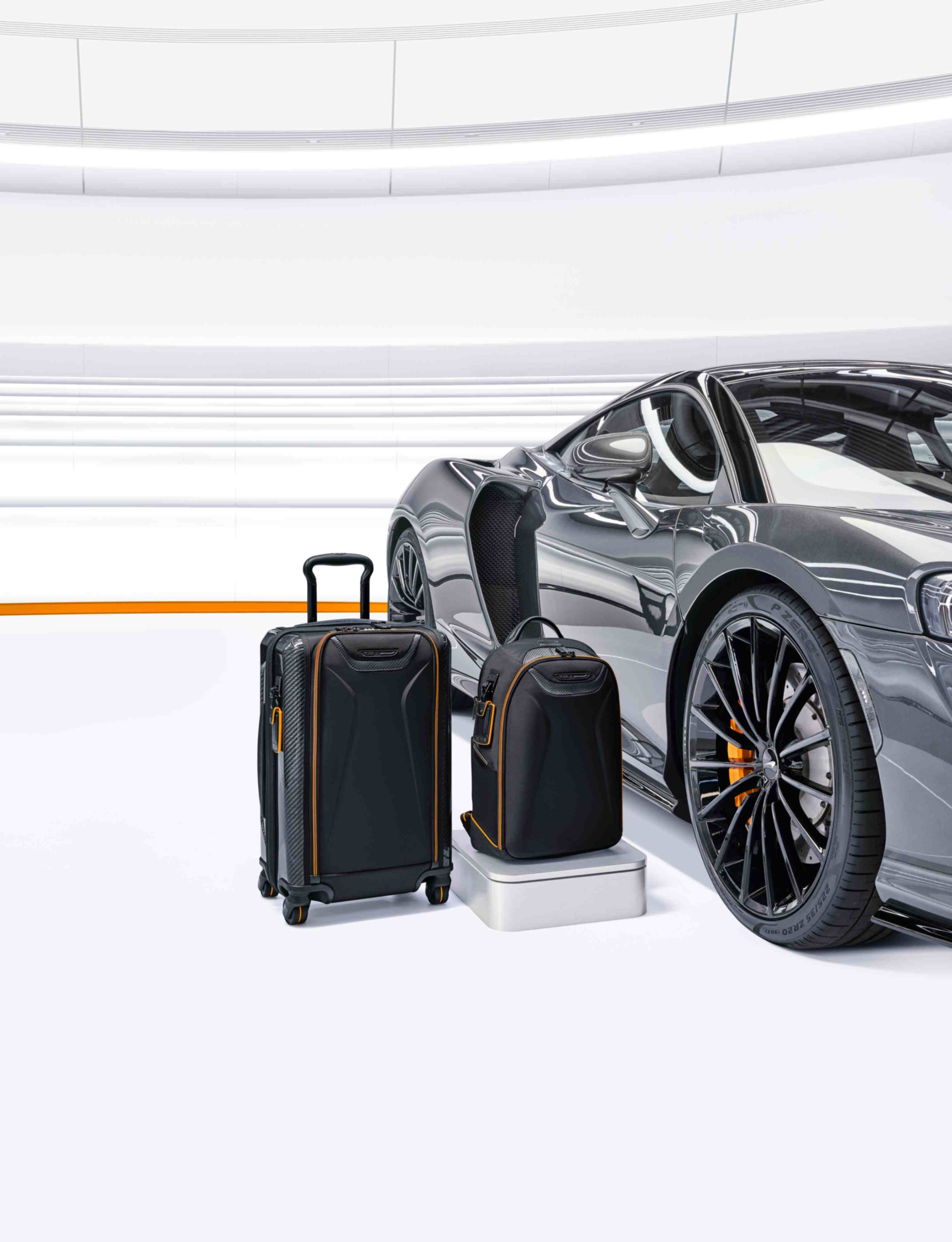 A TUMI | McLaren suitcase and backpack sat next to a grey McLaren in a white environment.