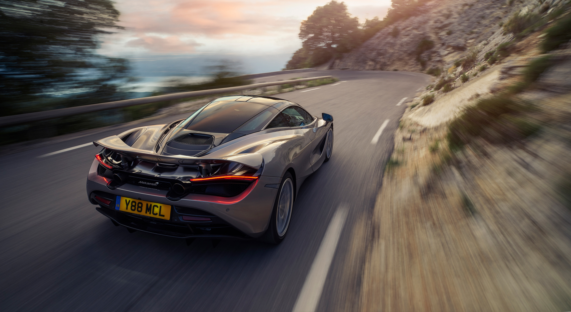 The rear of a McLaren 720S driving at high speed on a scenic road, with a motion blur effect.
