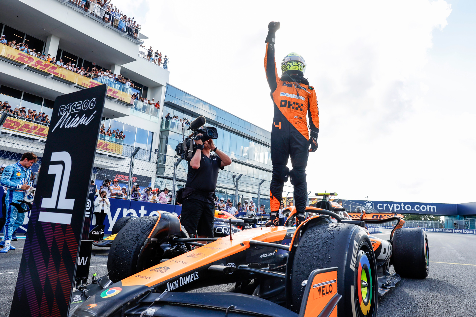 Lando Norris standing victoriously on his McLaren Formula 1 car after a face, raising his fist, still wearing his helmet, with a crowd and camera crew visible.