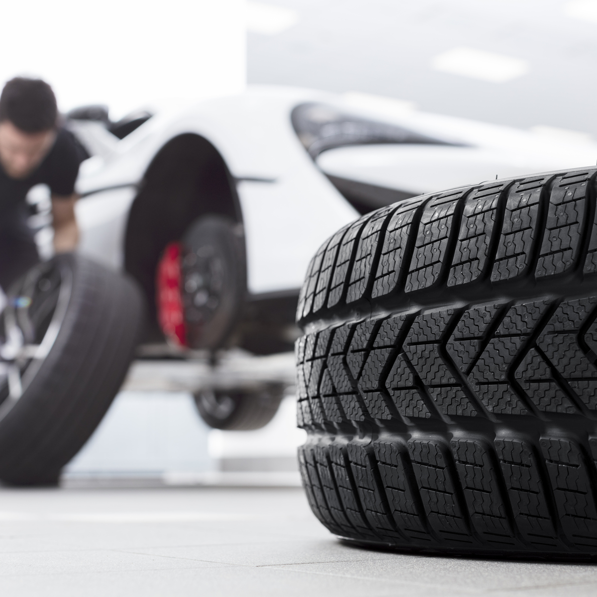 A large tire in the foreground with a McLaren technician changing a wheel on a white supercar in the background.