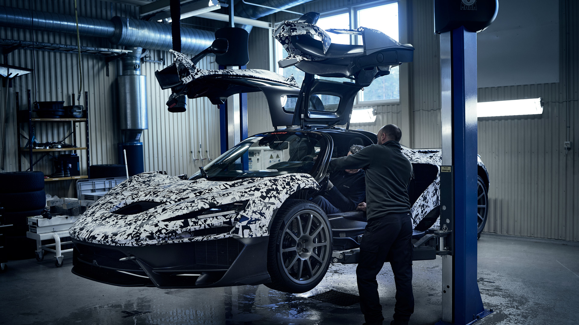 A camouflage-wrapped McLaren W1 prototype with its dihedral doors open is lifted on a hoist in a garage, with a technician working inside.