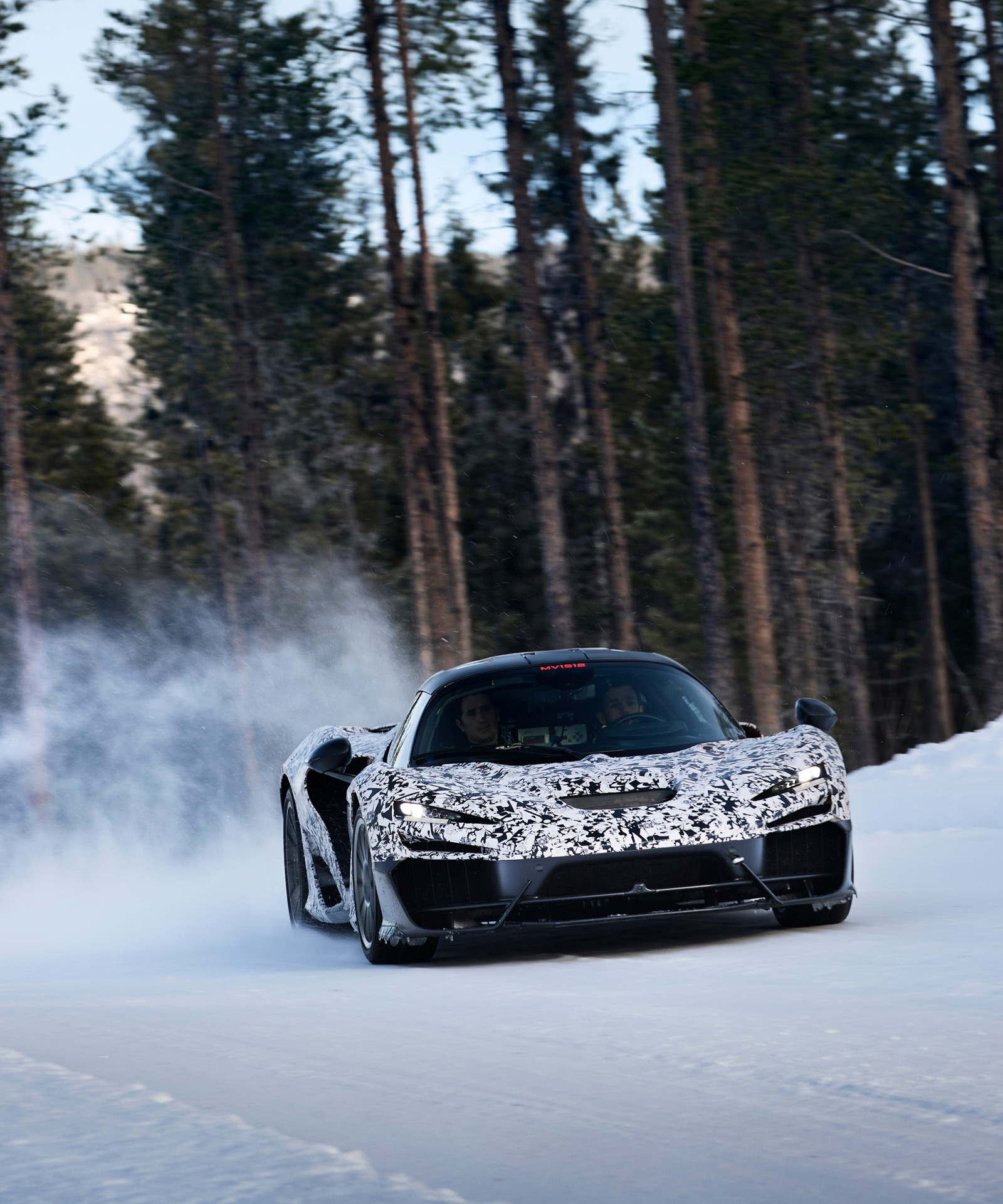 A camouflage-wrapped McLaren W1 prototype drifts through a snowy landscape with tall pine trees in the background, kicking up snow.