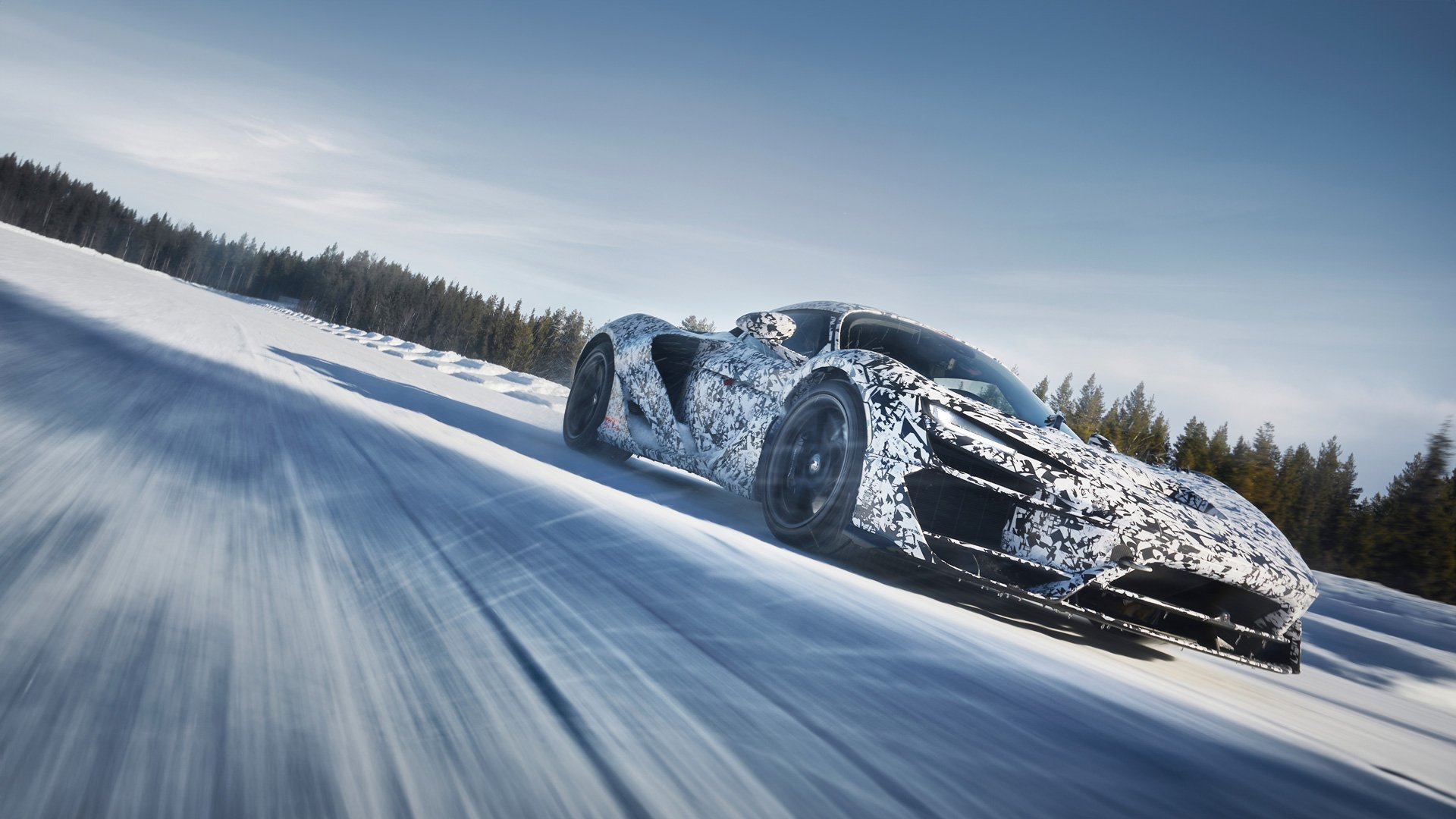 A McLaren W1 prototype covered in camouflage, blurring past on a snow-covered road with pine trees in the background, suggesting high-speed winter testing.