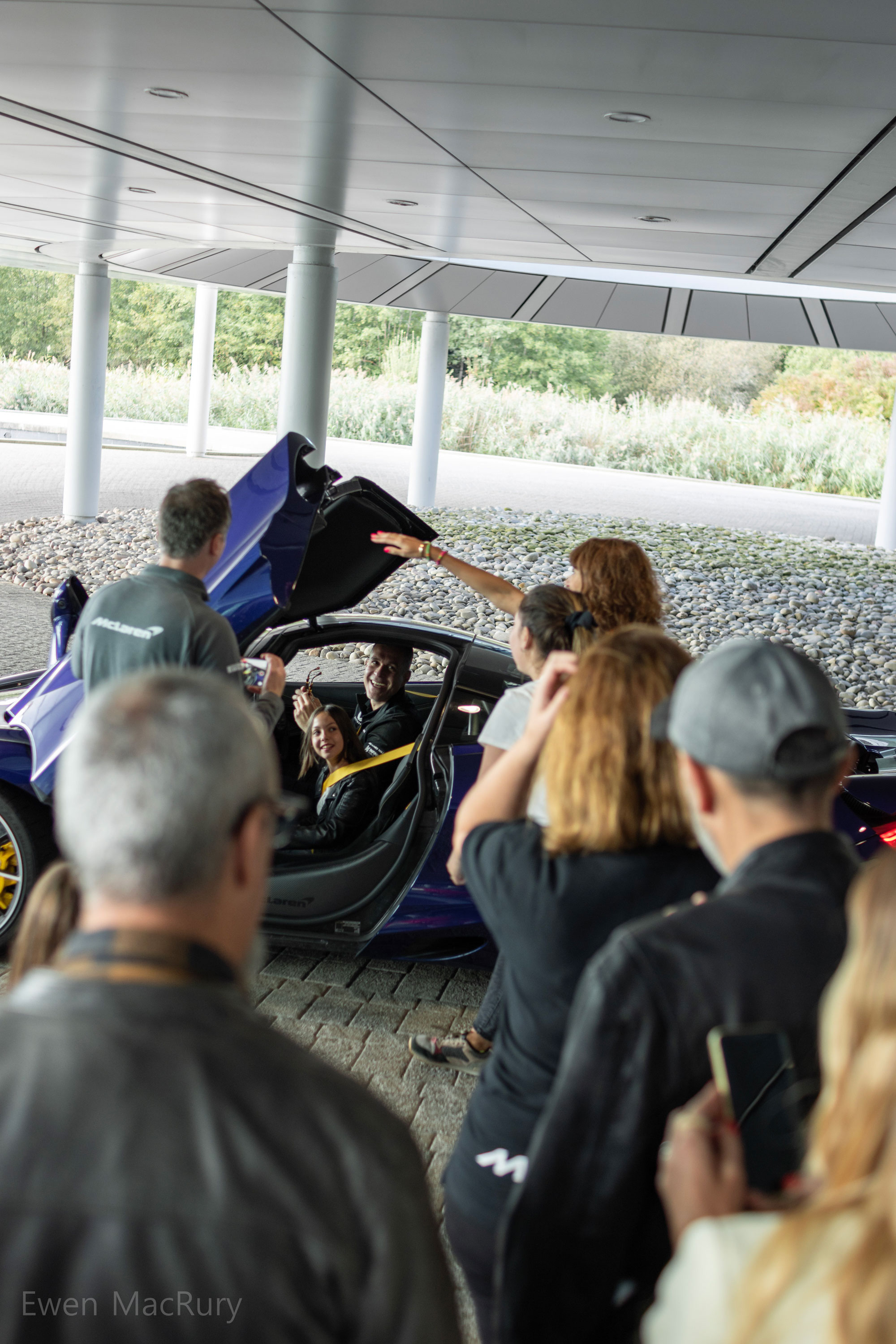 A group of people stood around a blue McLaren car. The car's door is open and two people are sitting in the car, both smiling at the camera.