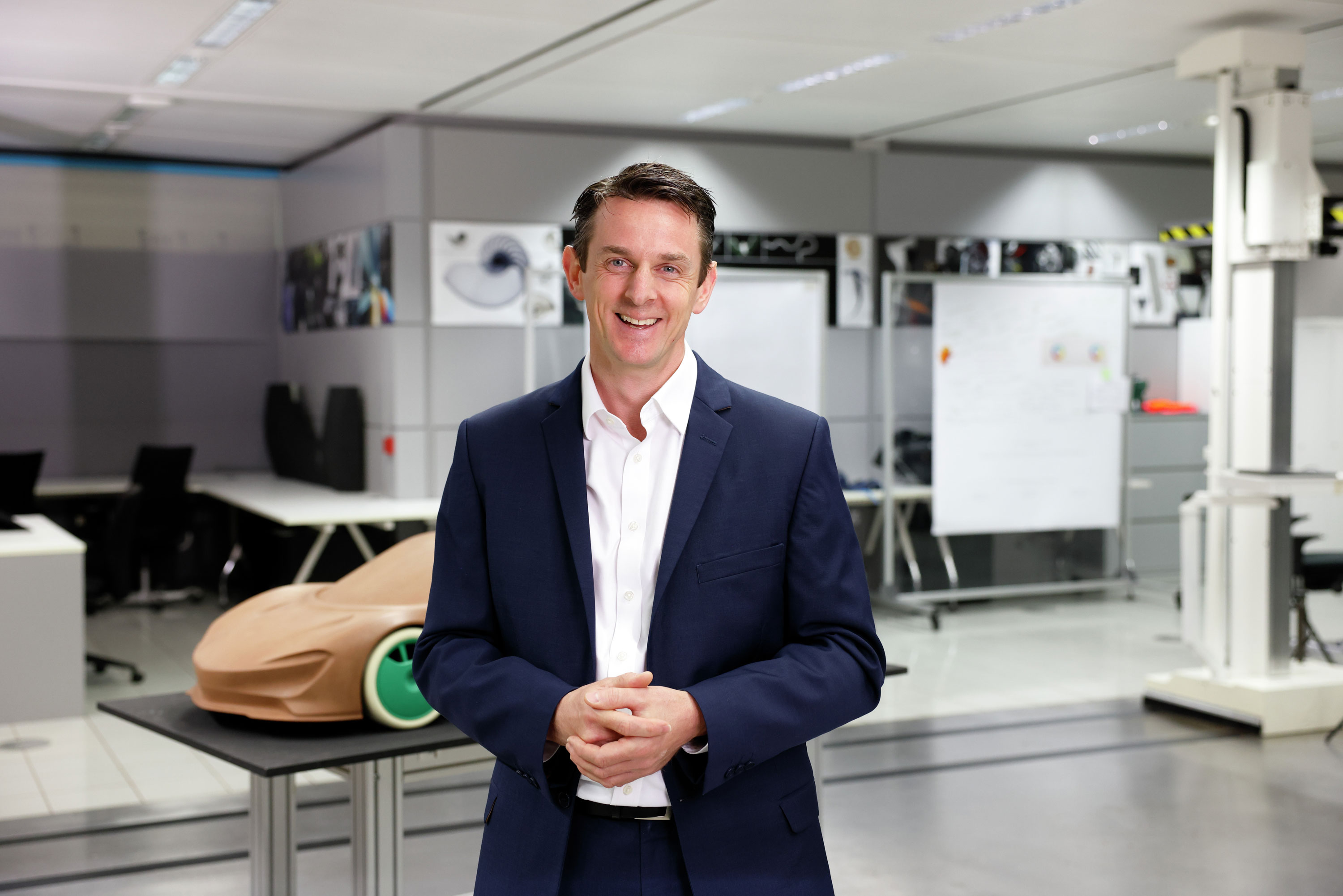 A smiling male McLaren employee in a suit, standing in a studio with a car model behind him.